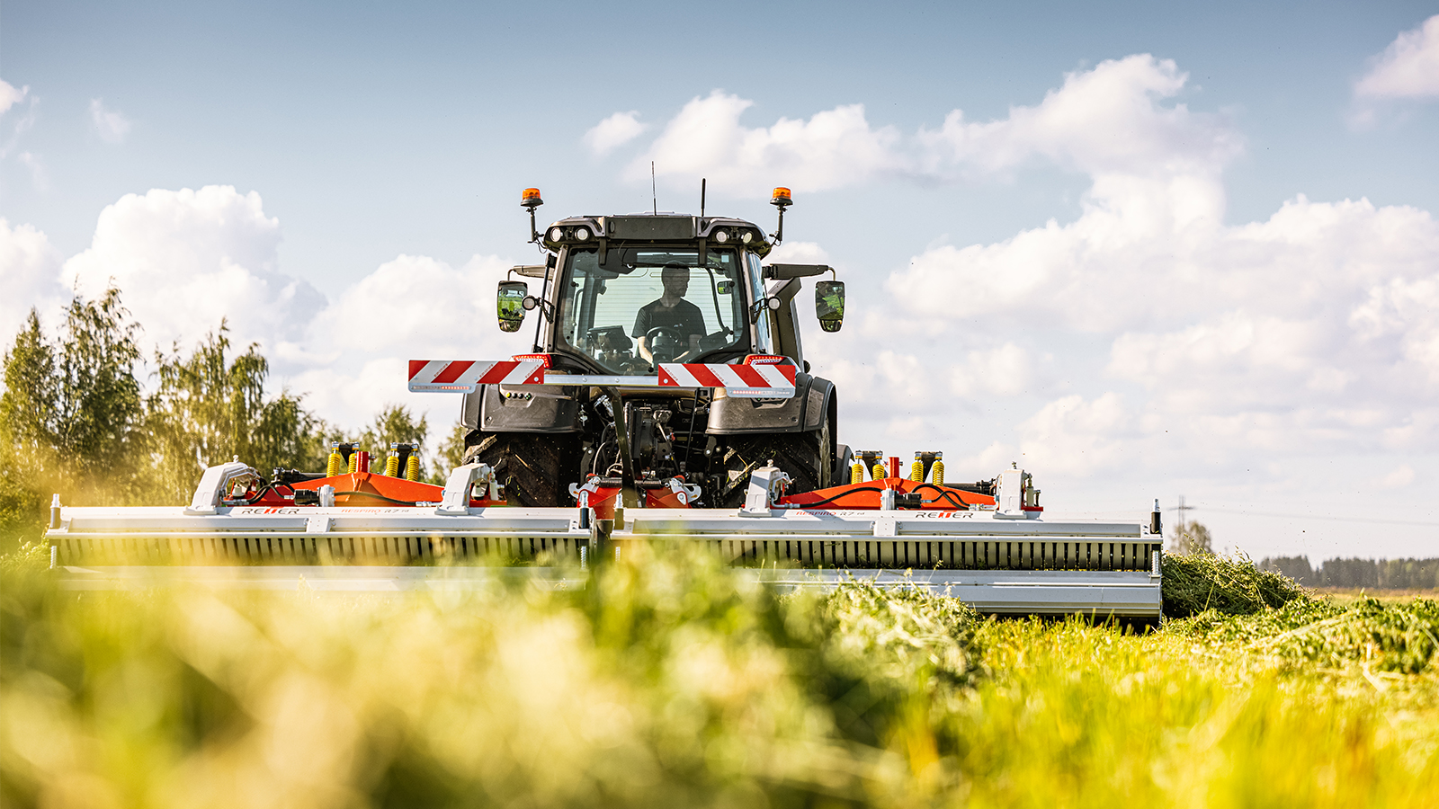 A Valtra T Series tractor with TwinTrac is mowing a lush green field with a wide front-mounted mower attachment. The machine is positioned centrally, driving toward the viewer through tall grass. Bright red and white safety markings stand out against the metallic equipment. A partly cloudy blue sky and distant tree line create a rural countryside backdrop. The overall scene emphasizes agricultural machinery, productivity, and open farmland.